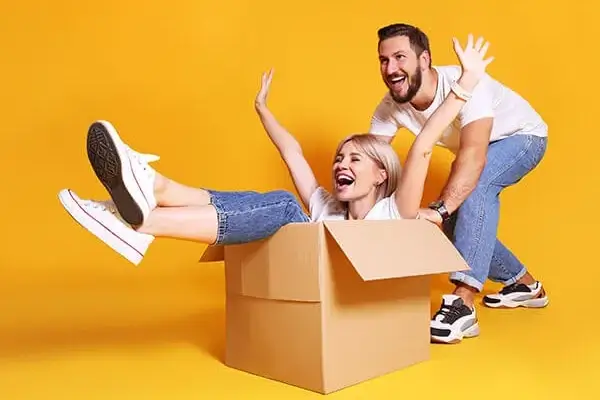 Joyful couple playing with cardboard box on yellow background.